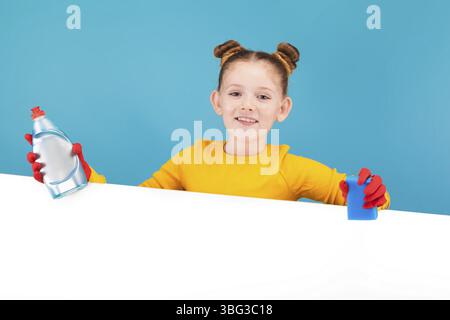 Isoliert auf blauem, entzückendem rothaarigem Mädchen mit Sommersprossen in Zitronenpullover und roten Latexhandschuhen, die Waschmittel und Schwamm über dem weißen Hintergrund halten Stockfoto