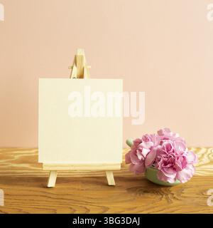 Staffelei und leere Leinwand mit Nelkenblumen auf Holztisch. Rosa Hintergrund Stockfoto
