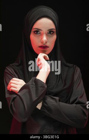 Porträt der schönen Ernsthafte junge muslimische Frau mit schwarzen Hijab als konservativ Fashion Concept mit der Hand in der Nähe von ihrem Gesicht auf schwarzem Hintergrund Stockfoto