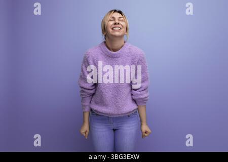 Porträt einer fröhlichen, süßen blonden Frau in einem Lavendelpullover auf einem lila Hintergrund mit Kopierraum Stockfoto