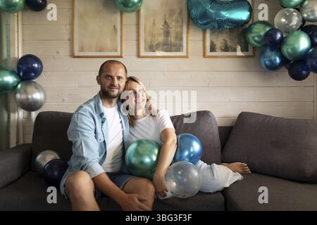 Ein glückliches Paar sitzt auf einer bequemen Couch, umgeben von blauen und grünen Ballons, und genießt ihre besondere Feier. Die Atmosphäre ist festlich und in Stockfoto