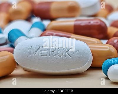 Weiße Pille mit MERCK-Aufdruck auf Tablettenstapel und farbenfrohe Kapseln in pharmazeutischer Optik. Stockfoto