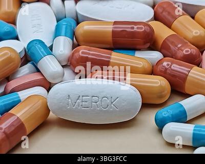 Weiße Pille mit MERCK-Aufdruck auf Tablettenstapel und farbenfrohe Kapseln in pharmazeutischer Optik. Stockfoto