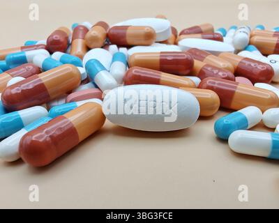 Weiße Pille mit MERCK-Aufdruck auf Tablettenstapel und farbenfrohe Kapseln in pharmazeutischer Optik. Stockfoto