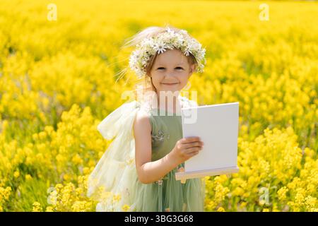 Fröhliches 5-Jähriges Mädchen mit Blumenkrone und Feenflügeln, das leere Leinwand auf hölzerner Staffelei hält und im Feld gelber Blumen posiert. Stockfoto