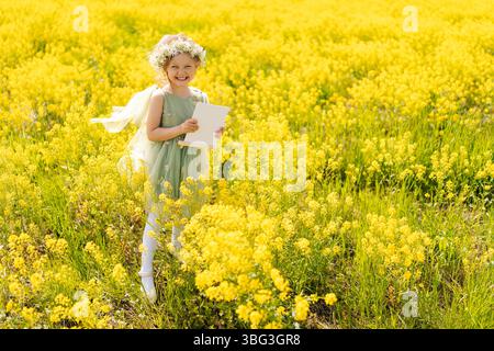 Porträt eines fröhlichen Mädchens in Feenkleidung und floralem Kopfschmuck, der mit leerem Leintuch inmitten einer goldenen Wildblumenwiese posiert, Sonnenlicht Stockfoto