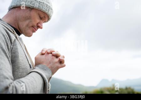 Erwachsener Mann, der auf einem Hügel steht und die Hände schließt, die Augen mit grauer Mütze und Kapuze schließt, Kopierraum Stockfoto