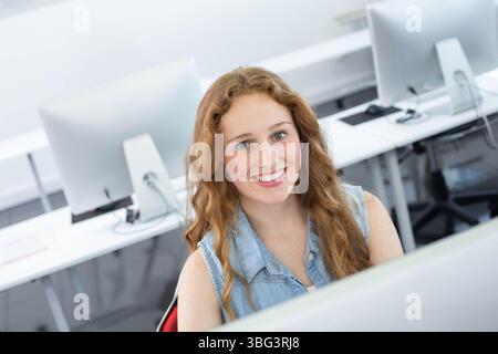Eine Schülerin lächelt und benutzt einen Desktop-Computer am weißen Schreibtisch im Computerlabor mit Denim-Hemd Stockfoto