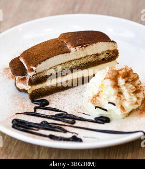 Tiramisu-Kuchen mit Schlagsahne und Schokoladensauce auf weißem Teller Stockfoto