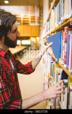 Der Mann schiebt ein blaues Hardcover-Buch aus dem Regal im Bibliotheksraum, während er ein rotes kariertes Hemd trägt Stockfoto