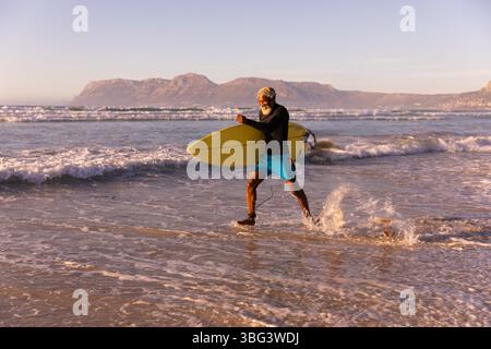 Ranghoher afroamerikanischer Mann, der am Strand durch das Wasser geht und Surfbrett in Boardshorts trägt Stockfoto