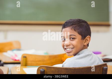 Asiatischer Junge, der am Holzschreibtisch sitzt, im Klassenzimmer auf Papieren unter Tafel schreibt, Kopierraum Stockfoto