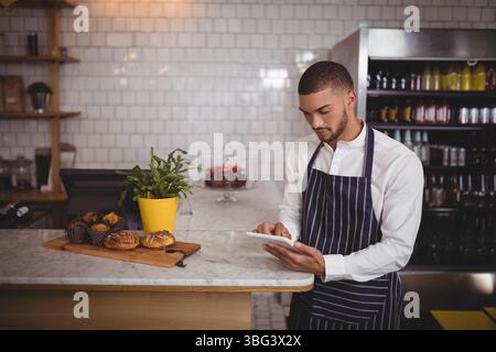 Mann, der gestreifte Schürze mit Tablet am Café-Theke mit Gebäck, Getränkespot, Kopierraum trägt Stockfoto