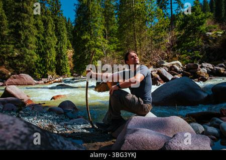 Mann Tourist sitzt auf Stein am Ufer des Flusses in den Bergen Stockfoto