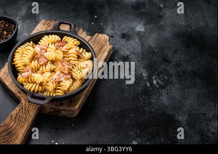 Fusilli Carbonara Nudeln mit knusprigen Speckstückchen und Parmesankäse in einer Pfanne. Schwarzer Hintergrund. Draufsicht. Stockfoto