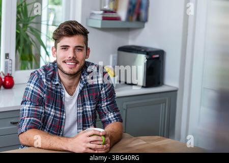 Mann, der am Küchentisch sitzt und einen Keramikbecher in der Nähe des Toasters hält, und eine Obstschale, die auf das Fenster blickt Stockfoto