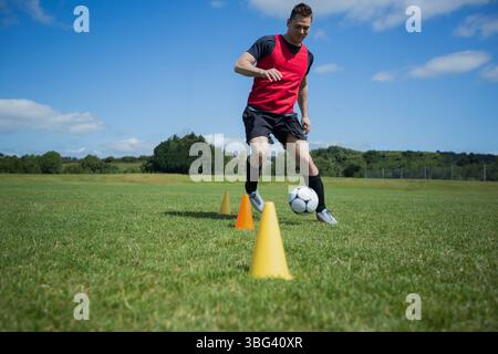 Männlicher Athlet dribbelt Fußball mit rotem Lätzchen und Stollen um Trainingskegel auf dem Rasenfeld Stockfoto
