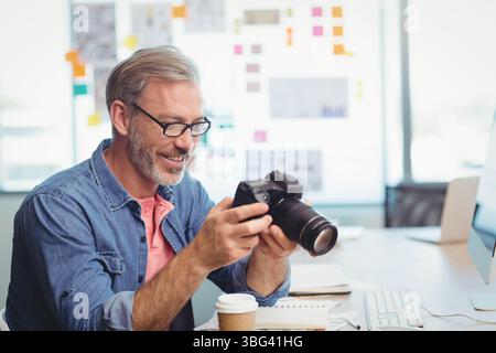 Mann mittleren Alters in Denim-Hemd, der die DSLR-Kamera am Schreibtisch mit Kaffeetasse und Notebook inspiziert Stockfoto