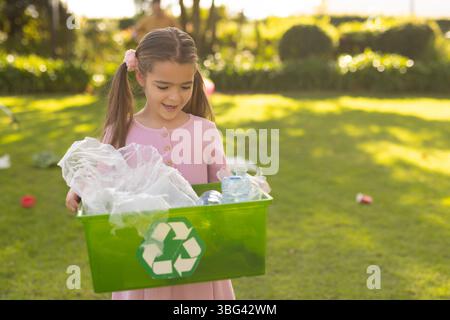 Mädchen in rosafarbenem Kleid, das auf Gras steht und einen grünen Recyclingbehälter mit Plastikflaschen und -Taschen hält Stockfoto