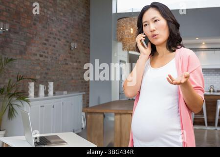 Schwangere asiatische Frau, die im Innenraum zu Hause steht und auf dem Smartphone per Laptop auf dem Tisch, Kopierraum anruft Stockfoto