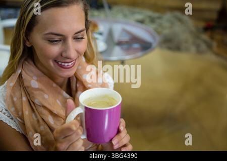 Frau, die am Couchtisch sitzt und einen pfirsichfarbenen Schal trägt, der zu Hause einen großen rosa Becher hält Stockfoto