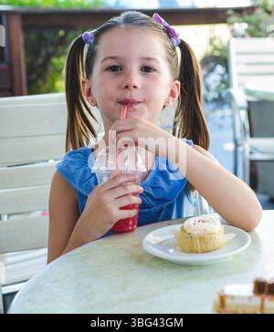 Mädchen, das auf der Terrasse des Cafés sitzt und knabbernd rotes Getränk durch Plastikbecher schlürft und Cupcake knabbert Stockfoto