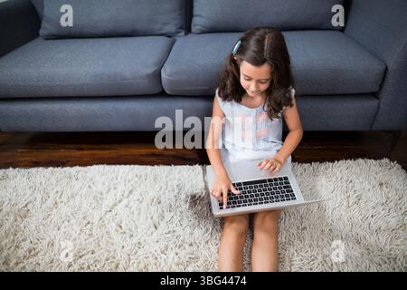 Mädchen, das auf weißem Teppich vor einem dunkelgrauen Sofa sitzt und auf silbernem Laptop arbeitet Stockfoto