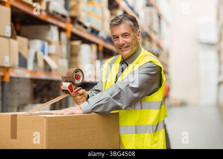 Mann mittleren Alters, der eine gut sichtbare Weste trägt, die einen Karton mit Klebeband in den Lagerregalen versiegelt Stockfoto