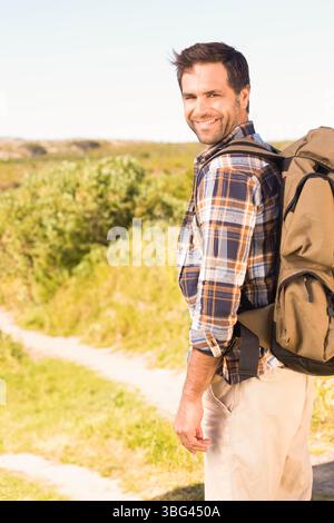 Olivgrüner Rucksack wird auf einem ländlichen Wanderweg getragen, der sich durch Sträucher unter blassblauem Himmel schlängelt Stockfoto