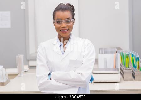 Afroamerikanische Frau, die auf der Laborbank steht, Laborkittel, Schutzbrille, Handschuhe in der Nähe von Reagenzglasgestell trägt Stockfoto