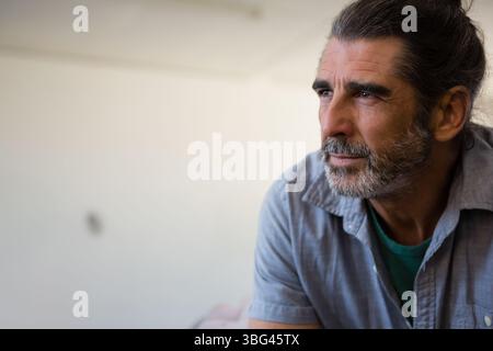 Reifer Mann sitzt im Studio zu Hause und blickt nach rechts an eine leere, weißweiße Wand, Kopierraum Stockfoto