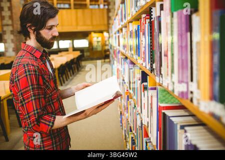 Mann, der im Büchergang steht und Buch in der Nähe von Bücherregalen, Lesetischen, Kopierraum hält Stockfoto
