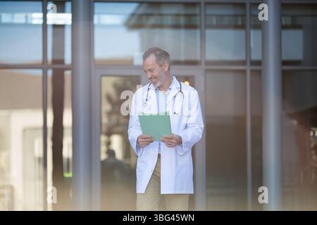Männlicher Arzt mittleren Alters, der am Eingang läuft, Laborkittel und Stethoskop trägt, grüne Mappe hält Stockfoto