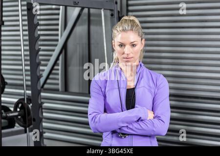 Frau, die mit gekreuzten Armen im Fitnessstudio vor einem Stahlträger mit Hantelplatten steht und eine Stoppuhr trägt Stockfoto