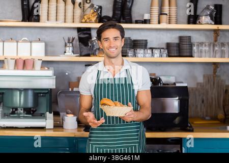 Männlicher Barista, mittelgroßer Erwachsener, trägt Schürze und hält einen Korb mit verschiedenen Brötchen an der Bäckerei Stockfoto