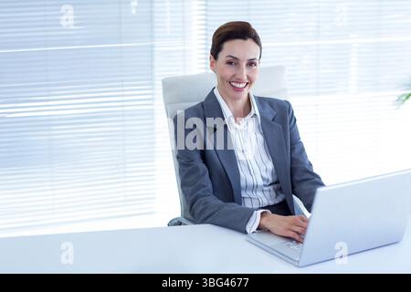Frau mit Aktivkohleblazer und Bluse, die auf silbernem Laptop am Schreibtisch in der Nähe von Fensterläden schreibt Stockfoto