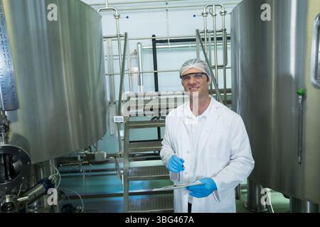 Männlicher Techniker in Schutzausrüstung, der Tanks in Verarbeitungsbetrieben mit Klemmbrett und Stift inspiziert Stockfoto