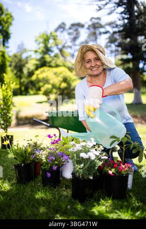 Ältere Frau, die im Garten kniet und Blumen mit grüner Gießkanne tränkt, mit Strohhut Stockfoto