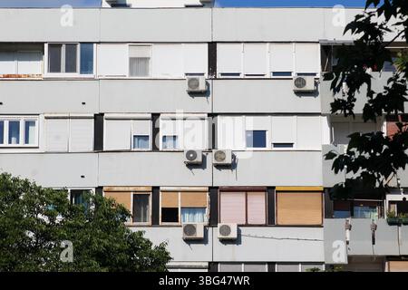 Fassade eines typischen sozialistischen Apartmentgebäudes mit einheitlichen Fenstern, geschlossenen Fensterläden und mehreren Klimaanlagen unter klarem blauem Himmel. Stockfoto