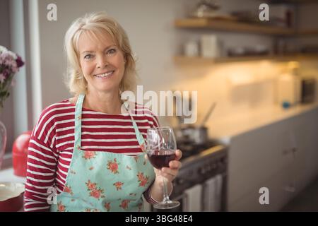 Ältere Frau, die in der Küche steht und Weinglas mit Stahlofen hält, mit Schürze Stockfoto