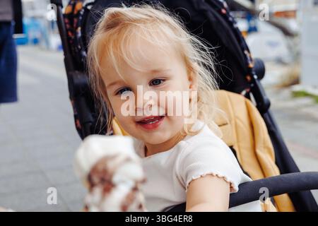 Ein glückliches zweijähriges blondes weißes Mädchen genießt ihre Vanilleeiskrem mit Ziegenmilch, während sie in einem Kinderwagen am Yachthafen sitzt. Reisen mit Stockfoto