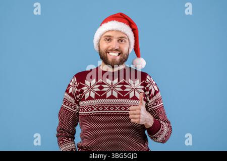 Positiver junger Mann in Urlaubspullover und Weihnachtsmütze, der Daumen hoch zeigt, lächelt und auf blauem Hintergrund auf die Kamera schaut Stockfoto