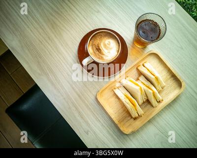 Eine gemütliche Frühstücksszene mit einer Tasse Kaffee mit Latte Art, einem Glas kaltem Getränk und einem Teller mit frischen Sandwiches auf einem Holztablett. Stockfoto