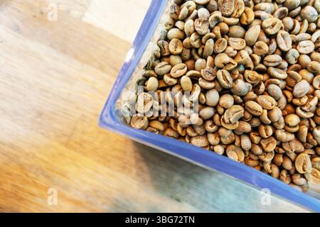 Eine Nahaufnahme der rohen Kaffeebohnen in einem durchsichtigen Behälter, die ihre erdige Textur vor einem hölzernen Hintergrund zeigen, perfekt für Speisen und Bevera Stockfoto