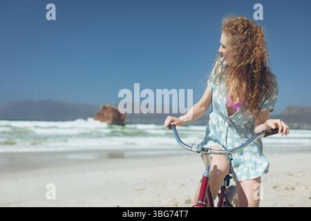 Frau radelt entlang der hellen Sandküste mit rotem Fahrrad in der Nähe von Felsvorsprüngen und Wellen Stockfoto