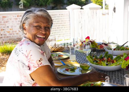 Hochrangige Afroamerikanerin lächelt, sitzt am Terrassentisch mit Salaten, Limonade, Kopierraum Stockfoto