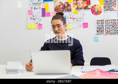 Asiatischer Mann, der auf silbernem Laptop am Schreibtisch mit bunten Haftnotizen und Strategietabellen tippt Stockfoto