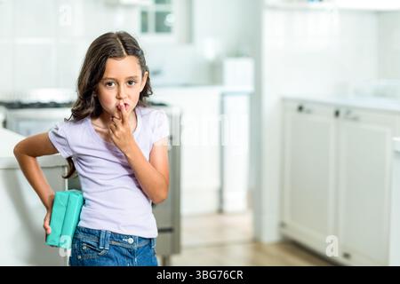 Mädchen, das in einer hellen, modernen Küche steht und türkisfarbene Geschenkbox hält und den Finger an die Lippen drückt Stockfoto