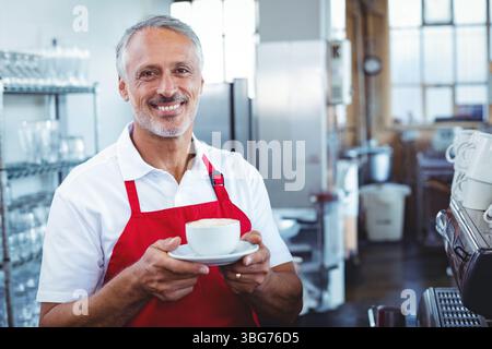 Ein älterer männlicher Barista, der hinter der Espressomaschine arbeitet und die Tasse auf einer Untertasse hält, trägt eine Schürze im Café Stockfoto