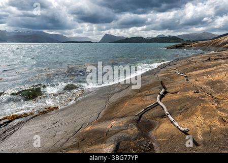 Wellen schlagen sanft gegen die zerklüfteten Felsen, während ein Stock am felsigen Strand unter einem bewölkten Himmel im Fjord liegt. Stockfoto